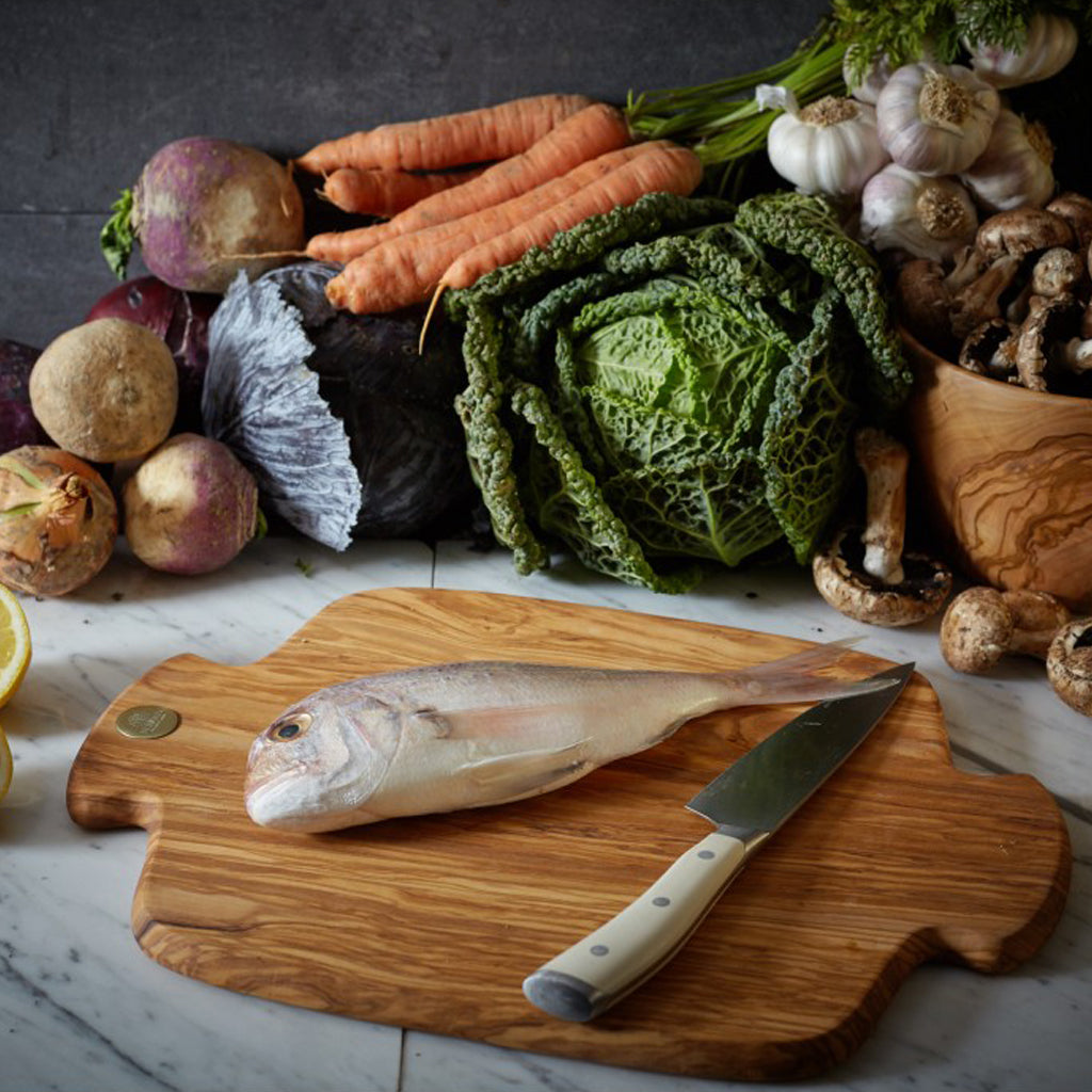 Berard Racine Large Olive Wood Cutting Board resting on a table surrounded by fresh vegetables.